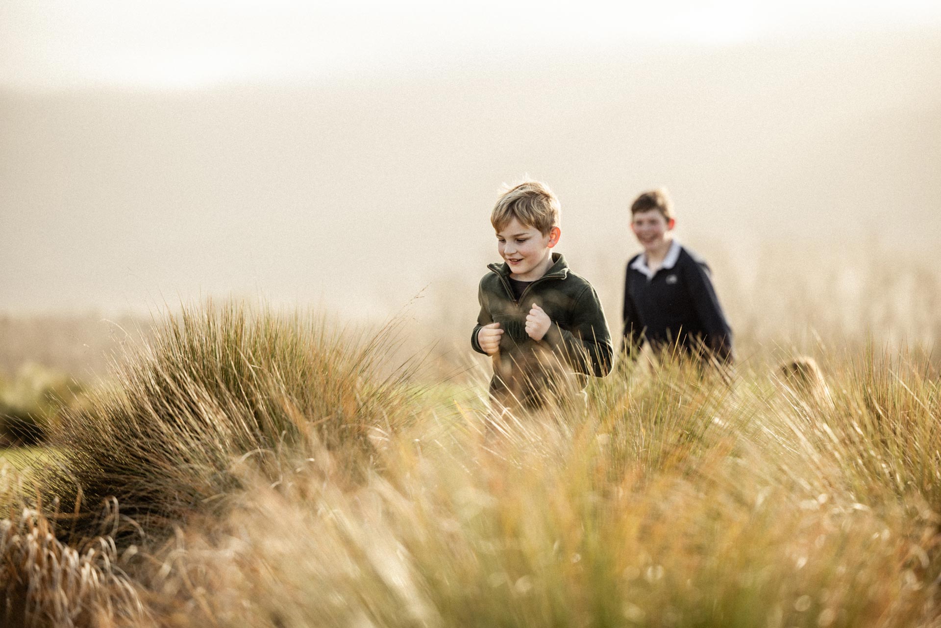 Two young boys running through the tussock grasses in the garden at Whakahā.